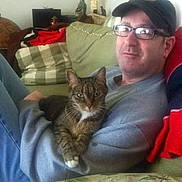 Phoebe is registered to the contest to win money with this photo: cat, tabby_cat, man, glasses, cap, couch, living_room, lap, pet, relaxed, sweater, pillow, blanket, indoor, home, furniture, portrait, smile, leisure, companion