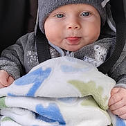 Leon is registered to the contest to win money with this photo: baby, child, infant, blanket, dinosaur_pattern, hat, gray_clothing, cute, tongue_out, smiling, seated, cozy, warm, portrait, face, hands, blue_eyes, indoor, plush, soft_texture