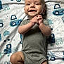 baby, smiling, onesie, grey_clothing, blanket, blue_pattern, construction_vehicles, infant, lying_down, hands_clasped, happy, cute, child, young_baby, indoor, soft_texture, toddler, portrait, playful, cheerful
