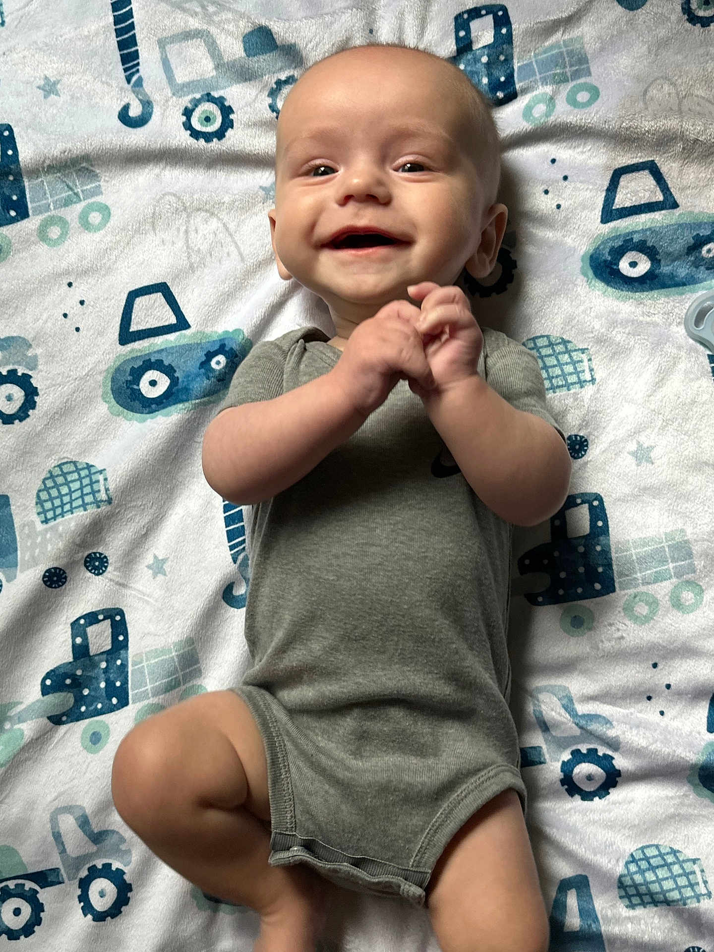 Jaxson joined the competition — help win amazing prizes! baby, smiling, onesie, grey_clothing, blanket, blue_pattern, construction_vehicles, infant, lying_down, hands_clasped, happy, cute, child, young_baby, indoor, soft_texture, toddler, portrait, playful, cheerful