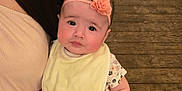 Leah is registered to the contest to win money with this photo: baby, child, headband, flower, bib, onesie, hand, person, adult, chair, wooden_floor, table, indoor, cute, looking, sitting, holding, pink, yellow, floral_pattern
