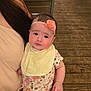 baby, child, headband, flower, bib, onesie, hand, person, adult, chair, wooden_floor, table, indoor, cute, looking, sitting, holding, pink, yellow, floral_pattern