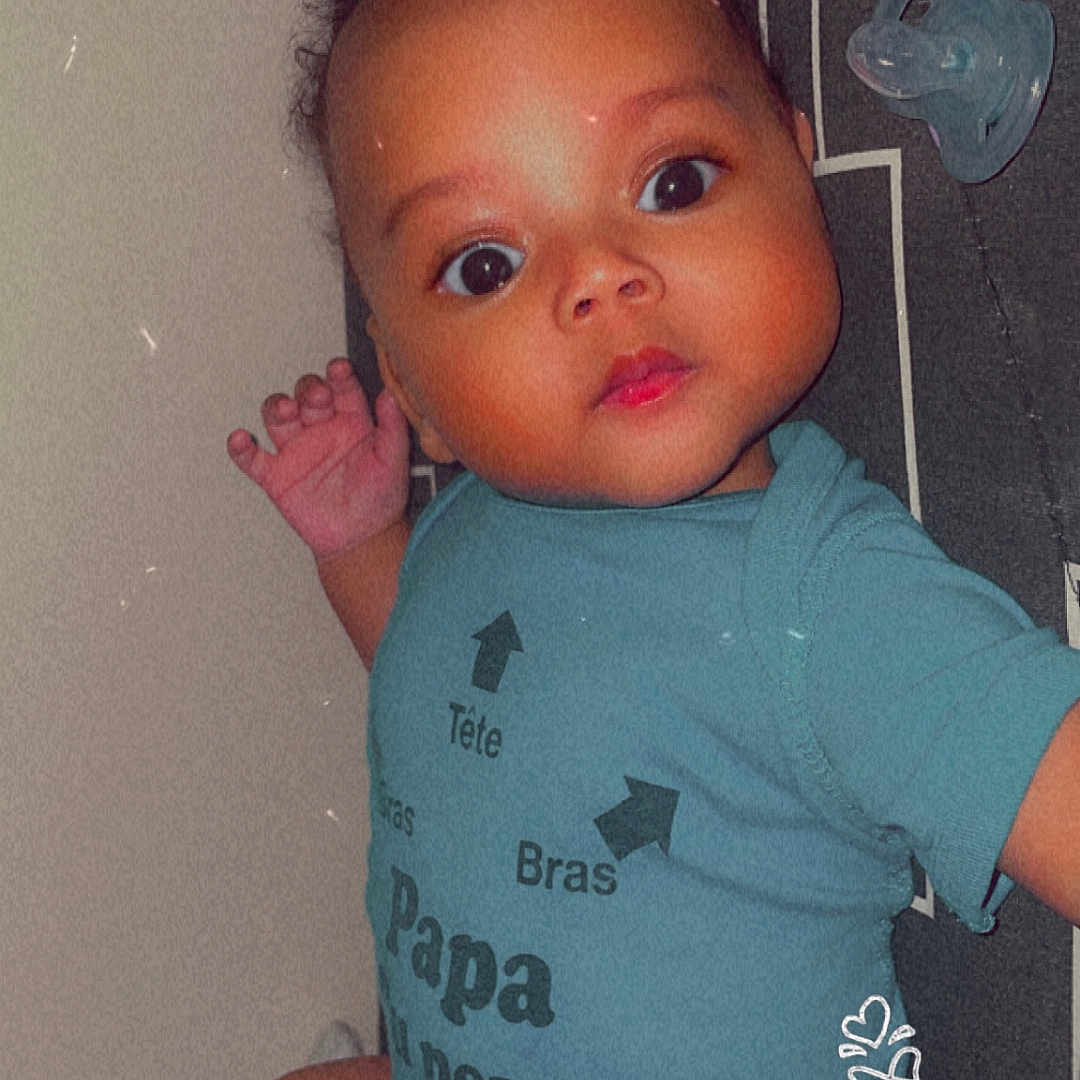 Heylann a rejoint le concours — aidez-le/la à gagner de superbes lots ! baby, blue_clothing, child, closeup, curly_hair, cute, expression, face, hand, indoors, infant, lying_down, onesie, pacifier, patterned_background, person, portrait, skin, soft_lighting, text_on_clothing