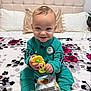 toddler, child, smiling, toy, board_books, bed, pillow, headboard, tracksuit, sneakers, indoor, happy, playing, seated, bedspread, floral_pattern, person, cute, young_child, casual_clothing