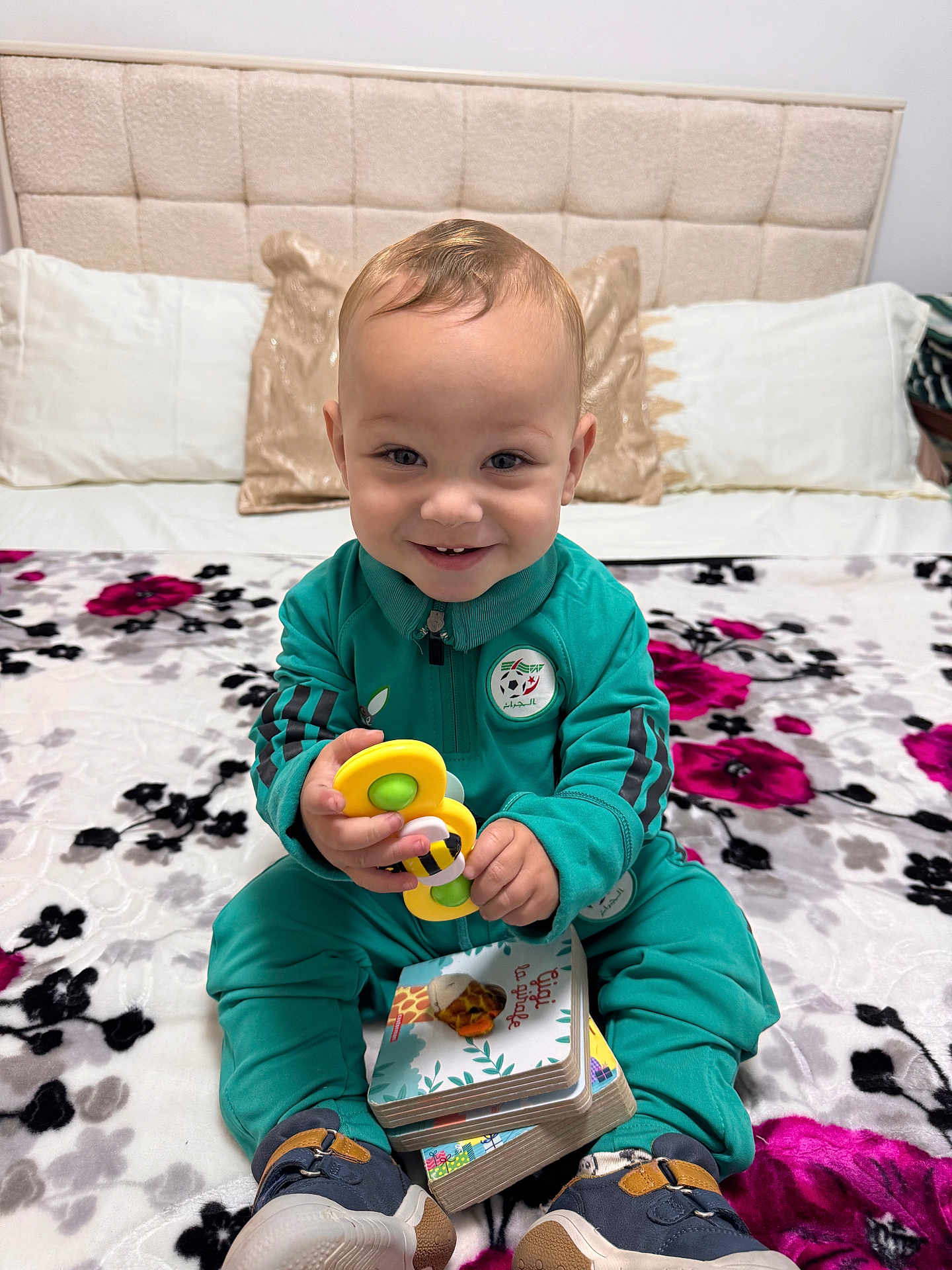 Ayden a rejoint le concours — aidez-le/la à gagner de superbes lots ! toddler, child, smiling, toy, board_books, bed, pillow, headboard, tracksuit, sneakers, indoor, happy, playing, seated, bedspread, floral_pattern, person, cute, young_child, casual_clothing