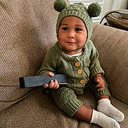 Hezekiah is registered to the contest to win money with this photo: baby, child, knit_hat, green_clothing, couch, remote_control, socks, indoor, cute, smiling, portrait, infant, warm_clothing, pom_poms, seated, home_interior, window_blinds, wooden_table, buttoned_sweater, happy