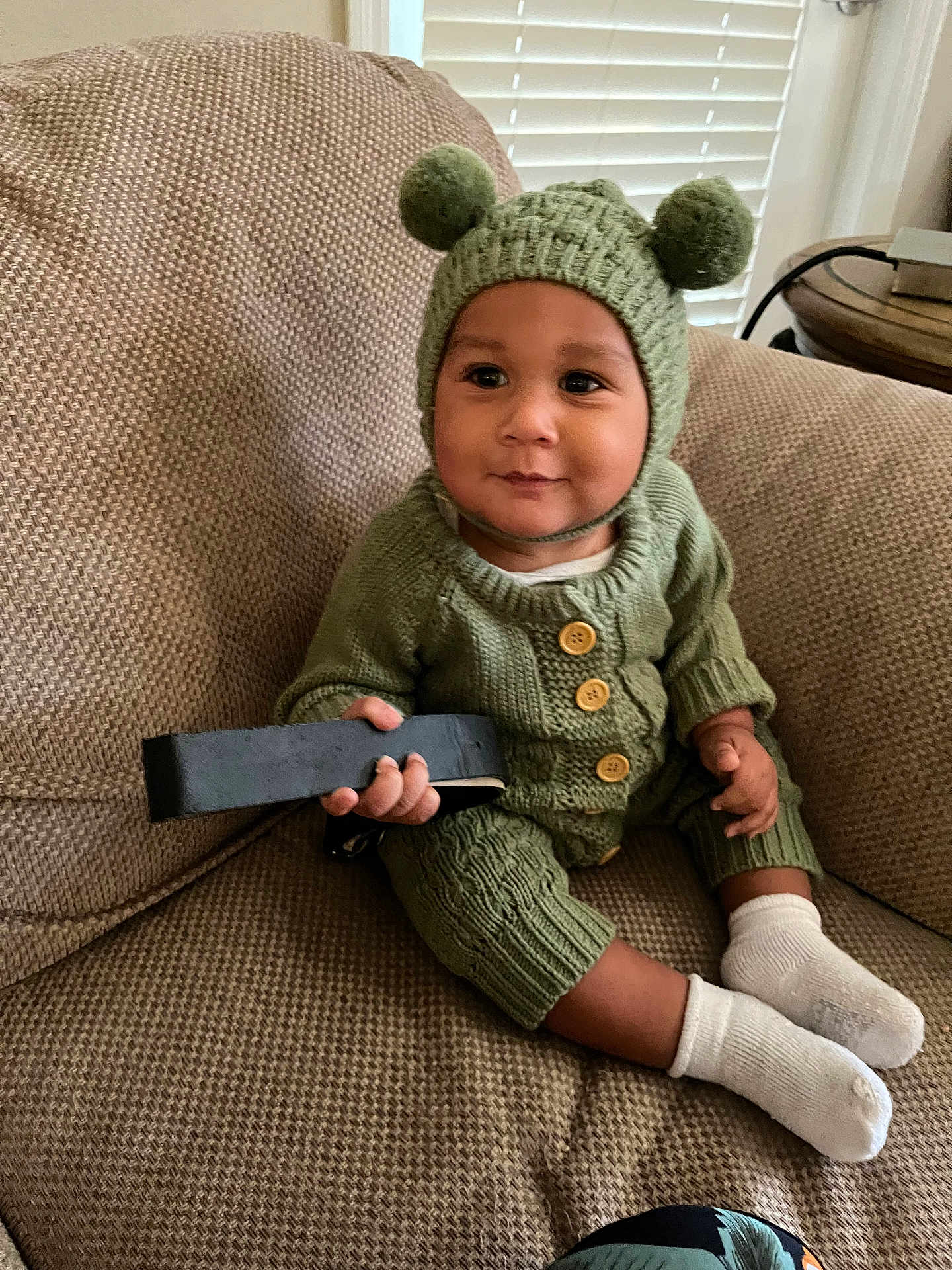 Hezekiah is registered to the contest to win money with this photo: baby, child, knit_hat, green_clothing, couch, remote_control, socks, indoor, cute, smiling, portrait, infant, warm_clothing, pom_poms, seated, home_interior, window_blinds, wooden_table, buttoned_sweater, happy
