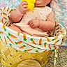 baby, infant, child, basket, carrot_pattern, rubber_duck, toy, smiling, happy, cute, sitting, pink_dress, quilt, carpet, indoor, portrait, face, hand, cheeks, young