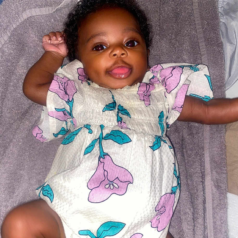 Raelyn is registered to the contest to win money with this photo: apparel, baby, bright, child, curly_hair, cute, face, floral_dress, happy, indoors, infant, lying_down, one_arm_up, portrait, skin, small_person, smiling, soft_surface, towel, young_child