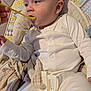 baby, feeding, spoon, pajamas, cushion, baby_seat, child, infant, face, hand, blanket, white_clothing, blue_eyes, indoor, closeup, person, food, comfort, soft, care
