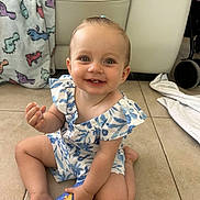 Nova-May is registered to the contest to win money with this photo: baby, child, smiling, sitting, floor, bottle, blue, pony_tail, teeth, indoor, blanket, dinosaur_pattern, couch, pillows, tile_floor, happy, cute, clothing, person, young_child