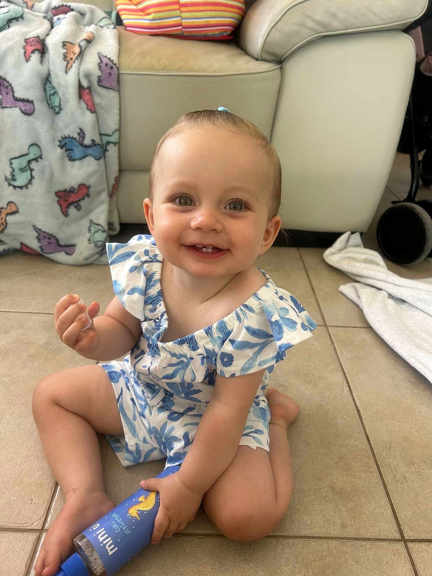 Nova-May is registered to the contest to win money with this photo: baby, child, smiling, sitting, floor, bottle, blue, pony_tail, teeth, indoor, blanket, dinosaur_pattern, couch, pillows, tile_floor, happy, cute, clothing, person, young_child