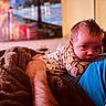 baby, adult, blanket, patterned_clothing, indoor, portrait, resting, curious, face, hand, colorful_background, painting, furniture, warm_lighting, cozy, person, head, skin, blue_shirt, soft_texture