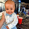 baby, child, smiling, bow_tie, shirt, blue_pants, hand, table, red_cup, smartphone, cafe, indoor, person, face, happy, portrait, seated, clothing, background, furniture