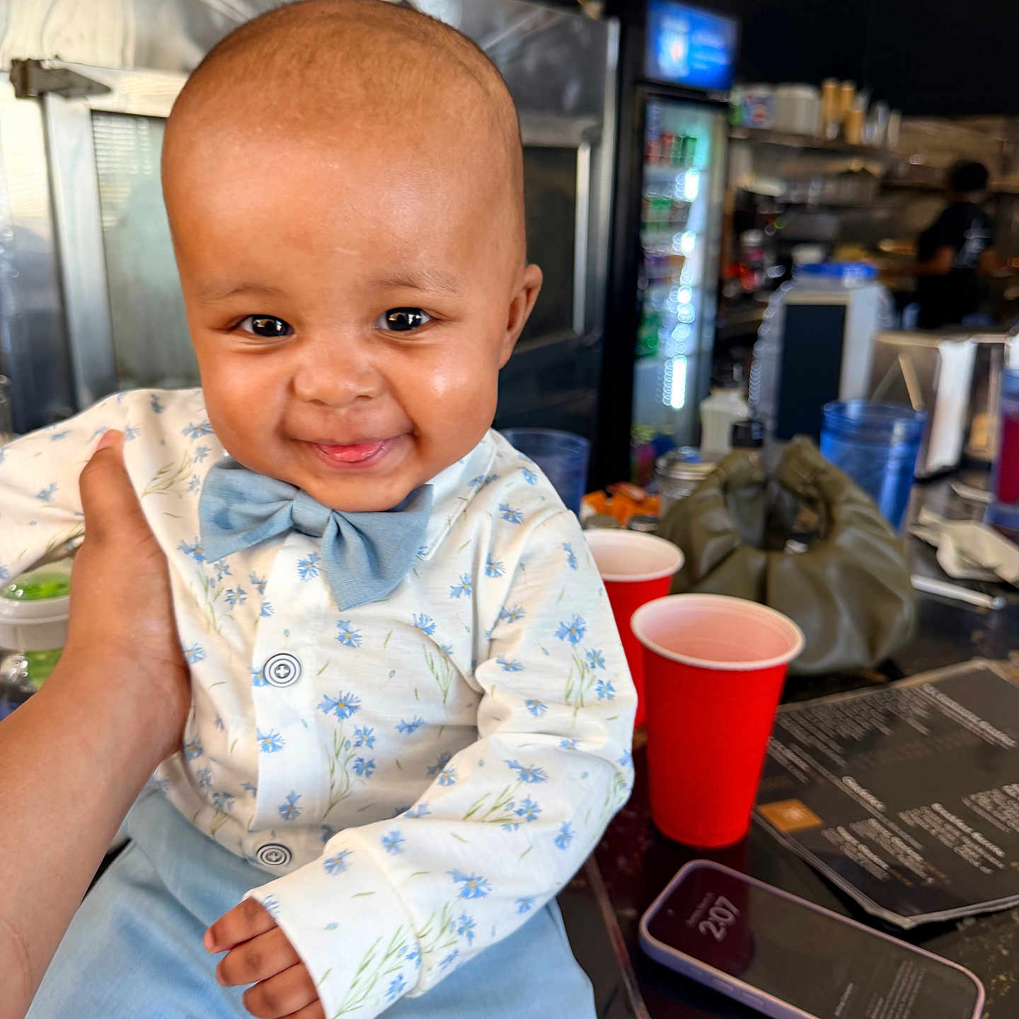 Jamarion is registered to the contest to win money with this photo: baby, child, smiling, bow_tie, shirt, blue_pants, hand, table, red_cup, smartphone, cafe, indoor, person, face, happy, portrait, seated, clothing, background, furniture
