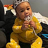 baby, child, tiara, wand, yellow_onesie, sitting, indoor, cute, face, hands, feet, black_background, plush_clothing, sparkle, accessory, portrait, curious, infant, young_child, chewing