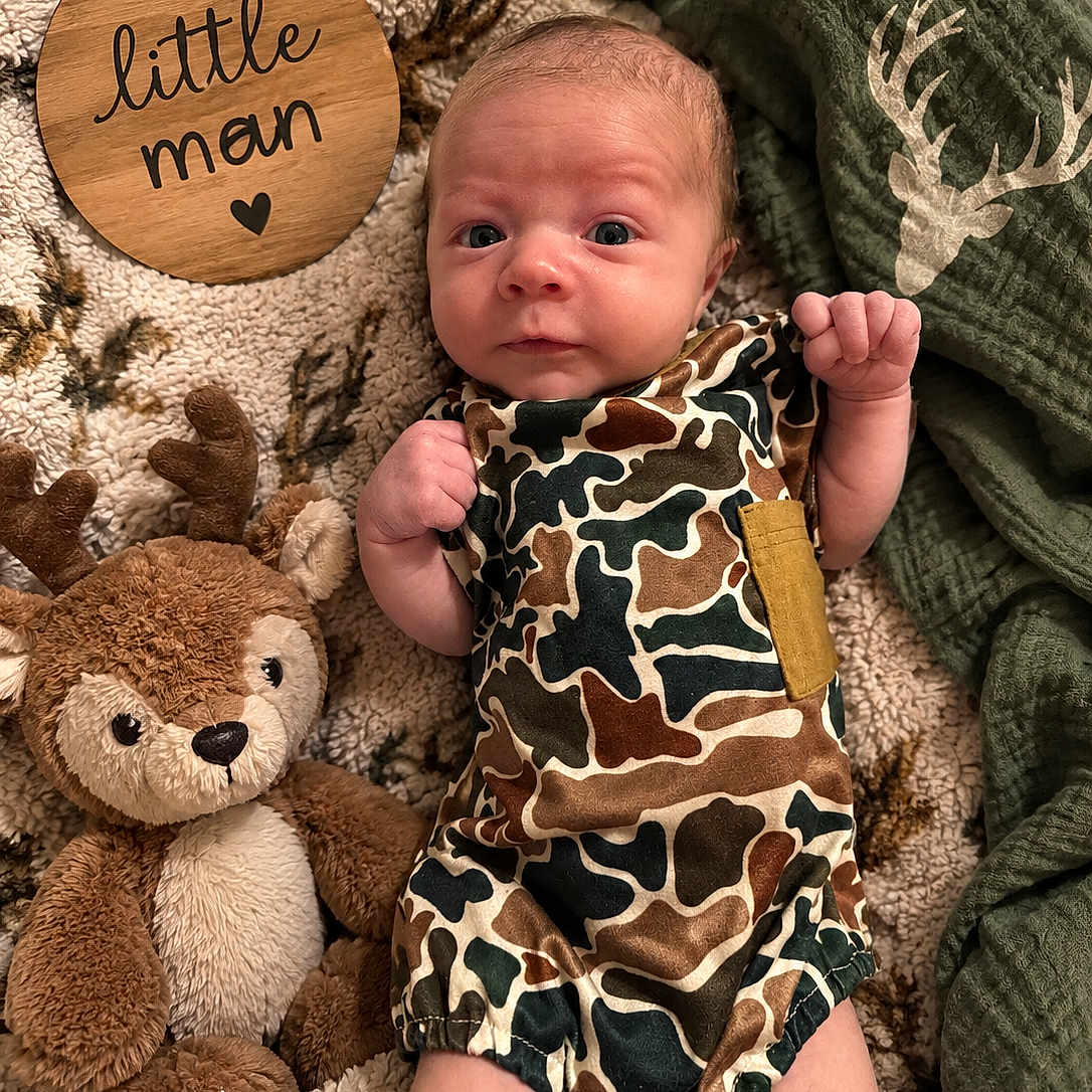 Sutton joined the competition — help win amazing prizes! baby, newborn, child, infant, portrait, lying_down, wide_eyed, face, smiling, camouflage, overalls, plush_toy, reindeer, stuffed_animal, blanket, wooden_sign, cozy, indoors, nursery, photography