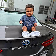 André joined the competition — help win amazing prizes! baby, child, car, toyota, camry, trunk, outdoor, parking_lot, blue_clothing, necklace, cross, smile, curly_hair, sneakers, vehicle, license_plate_area, rear_light, urban, person, happy