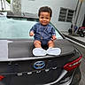 baby, child, car, toyota, camry, trunk, outdoor, parking_lot, blue_clothing, necklace, cross, smile, curly_hair, sneakers, vehicle, license_plate_area, rear_light, urban, person, happy