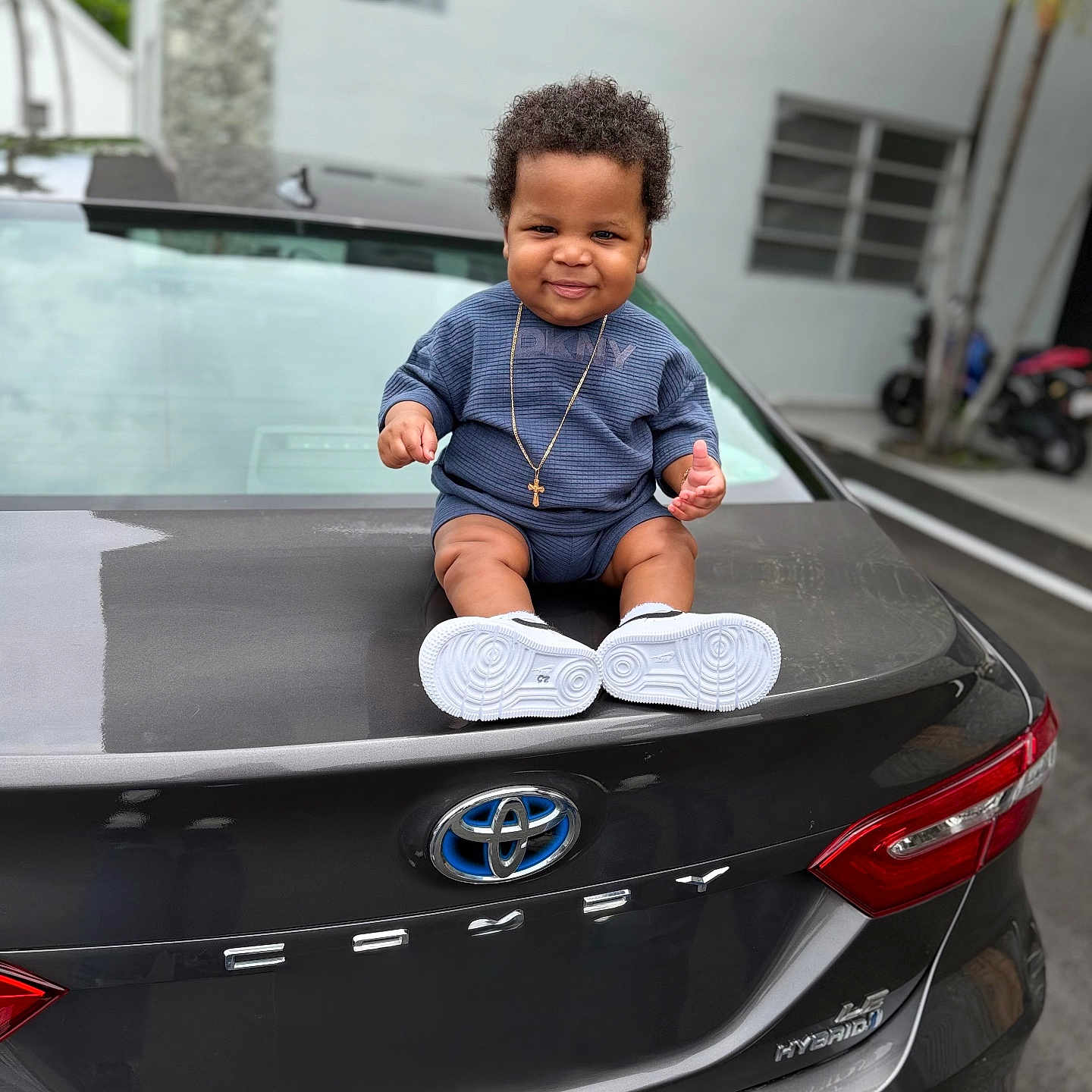 André joined the competition — help win amazing prizes! baby, child, car, toyota, camry, trunk, outdoor, parking_lot, blue_clothing, necklace, cross, smile, curly_hair, sneakers, vehicle, license_plate_area, rear_light, urban, person, happy