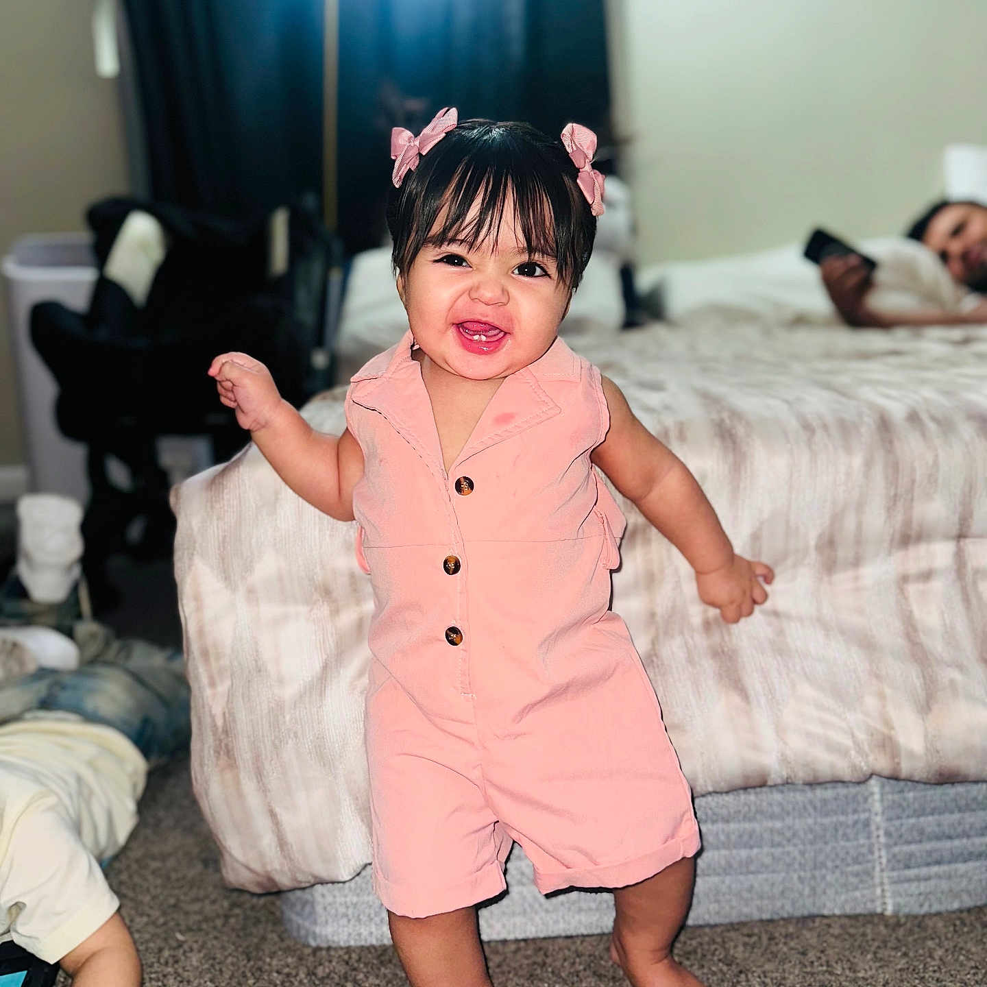 Kalye is registered to the contest to win money with this photo: bed, bedroom, bows, carpet, child, child_playing, cute, family, furniture, hair_bangs, happy, home, indoor, lamp, person, phone, pink_romper, smiling, standing, toddler