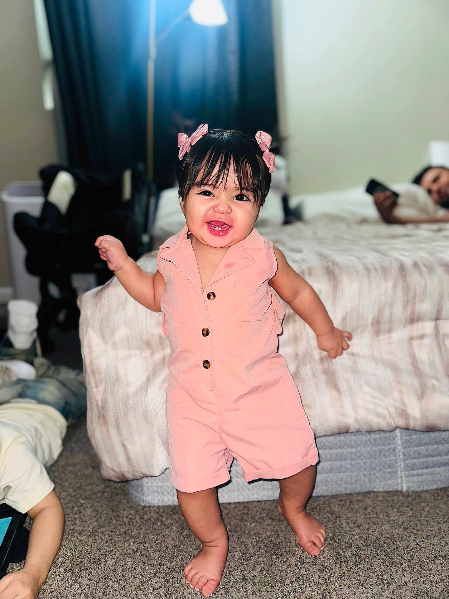 Kalye is registered to the contest to win money with this photo: bed, bedroom, bows, carpet, child, child_playing, cute, family, furniture, hair_bangs, happy, home, indoor, lamp, person, phone, pink_romper, smiling, standing, toddler