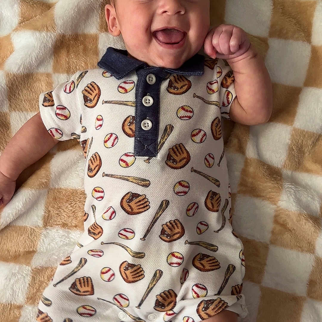 Gentry is registered to the contest to win money with this photo: baby, baseball, blanket, child, clothing, cute, fabric, fist, happy, indoor, infant, lying_down, pattern, person, portrait, shoes, smiling, socks, soft, sportswear