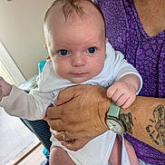 Oakley joined the competition — help win amazing prizes! baby, infant, person, adult, hand, tattoo, watch, clothing, white_onesie, blue_eyes, face, indoor, portrait, holding, sitting, furniture, purple_shirt, skin, expression, home