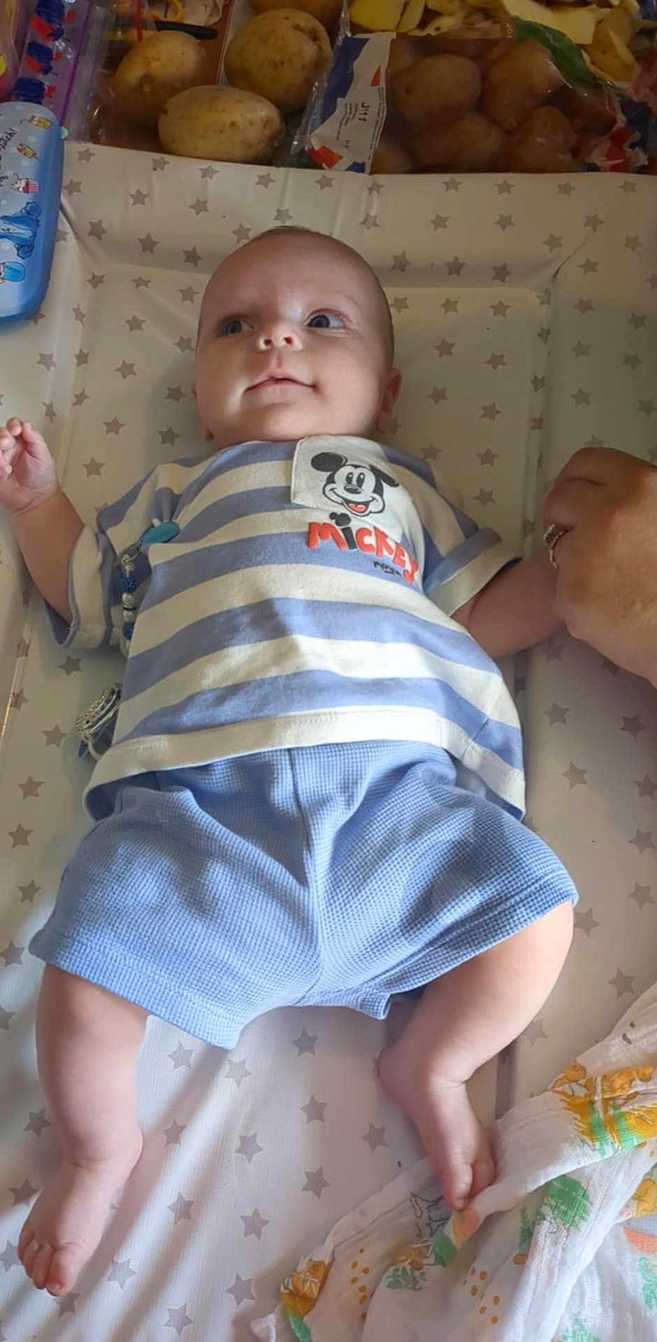 Oakley is registered to the contest to win money with this photo: baby, infant, child, mickey_mouse, striped_shirt, blue_shorts, star_pattern, hand_holding, potatoes, blanket, lying_down, cute, indoor, person, clothing, face, legs, foot, expression, toy
