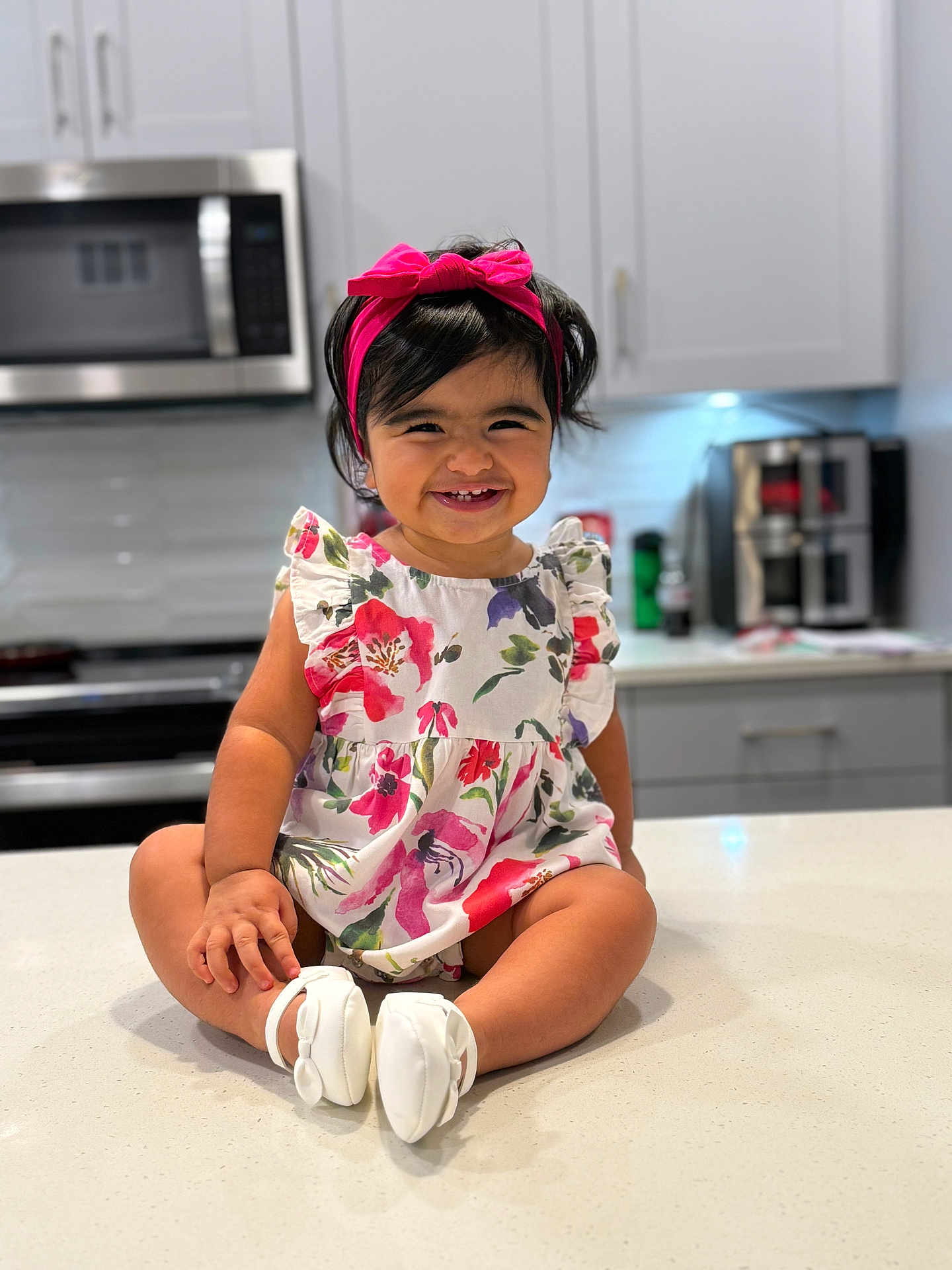 Amely joined the competition — help win amazing prizes! toddler, child, smiling, headband, pink, dress, floral, kitchen, counter, appliances, microwave, cabinets, modern, happy, sitting, white_shoes, indoors, person, cute, portrait