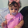baby, infant, child, portrait, sitting, pink_clothing, headband, bow, pacifier, pacifier_clip, wide_eyes, curly_hair, indoors, couch, blanket, close_up, front_facing, cute, baby_clothing, hand