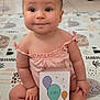 baby, balloons, child, cute, expression, floor, foot, hair_bow, hand, home, indoor, infant, milestone_card, patterned_mat, person, pink_outfit, play_mat, portrait, sitting, young_child