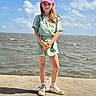 child, girl, pink_cap, smile, green_outfit, shorts, sneakers, socks, dock, water, sky, clouds, sunny, outdoor, standing, portrait, daylight, happy, fashion, casual
