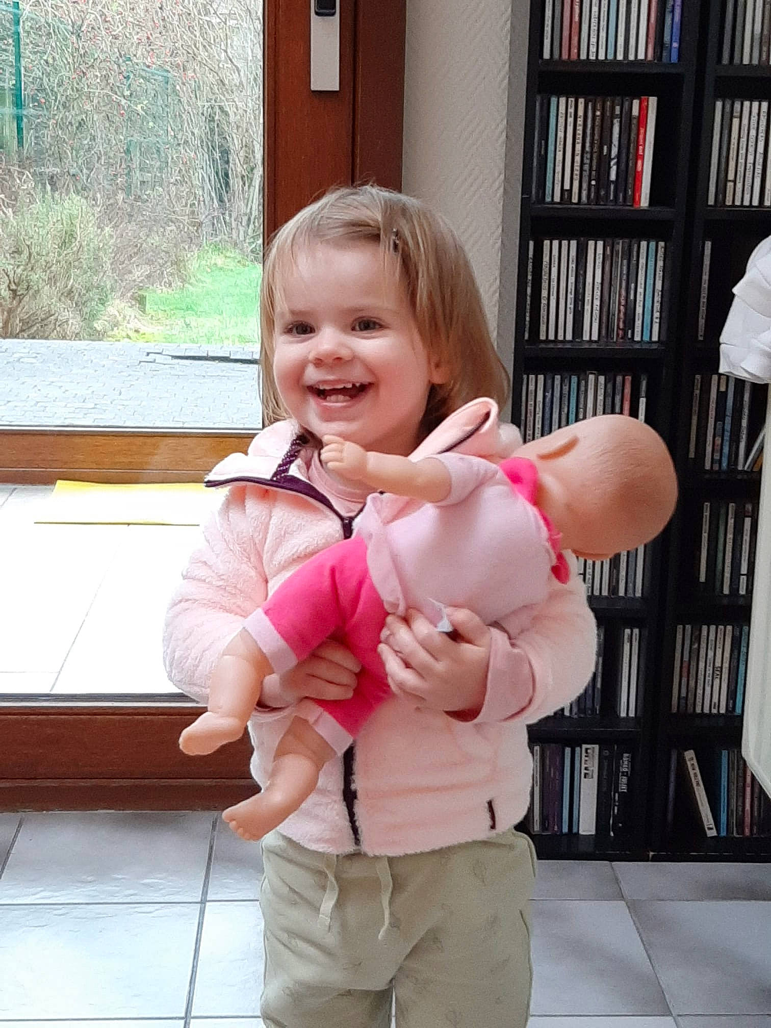 Melinda participe au concours pour gagner de l'argent avec cette photo : baby, baby_toddler_clothing, bookcase, cheek, eye, facial_expression, finger, gesture, happy, joint, joy, leg, person, pink, shelf, skin, sleeve, smile, standing, thumb