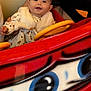 baby, child, person, ride, toy_car, red, yellow, white_clothing, fur_vest, cute, smiling, hand, arm, indoor, bright_colors, portrait, happy, head, face, seated