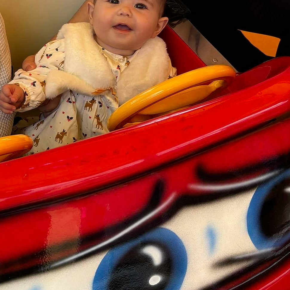 Taylana a rejoint le concours — aidez-le/la à gagner de superbes lots ! arm, baby, bright_colors, child, cute, face, fur_vest, hand, happy, head, indoor, person, portrait, red, ride, seated, smiling, toy_car, white_clothing, yellow
