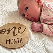 Blakely is registered to the contest to win money with this photo: baby, infant, one_month, blanket, wooden_sign, tongue_out, cute, newborn, floral_clothing, lying_down, close_up, skin, face, hand, milestone, soft_texture, indoor, portrait, child, young