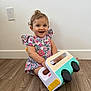 Ghevorchean Suzanna joined the competition — help win amazing prizes! toddler, child, toy, truck, floor, wooden_floor, curly_hair, floral_dress, smiling, happy, indoor, play, person, baby, cute, sitting, colorful, plastic_toy, joy, white_wall