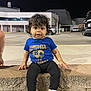 toddler, child, curly_hair, blue_tshirt, leggings, sneakers, stone_ledge, night, parking_lot, building, person, outdoor, casual_clothing, sitting, expression, urban, sidewalk, small_child, cute, curious