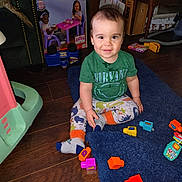 Aaron is registered to the contest to win money with this photo: casual_clothing, child, colorful, floor, furniture, green_shirt, happy, home, indoor, patterned_pants, person, playtime, rug, sitting, smiling, toddler, toy, toy_blocks, wooden_floor, young_child