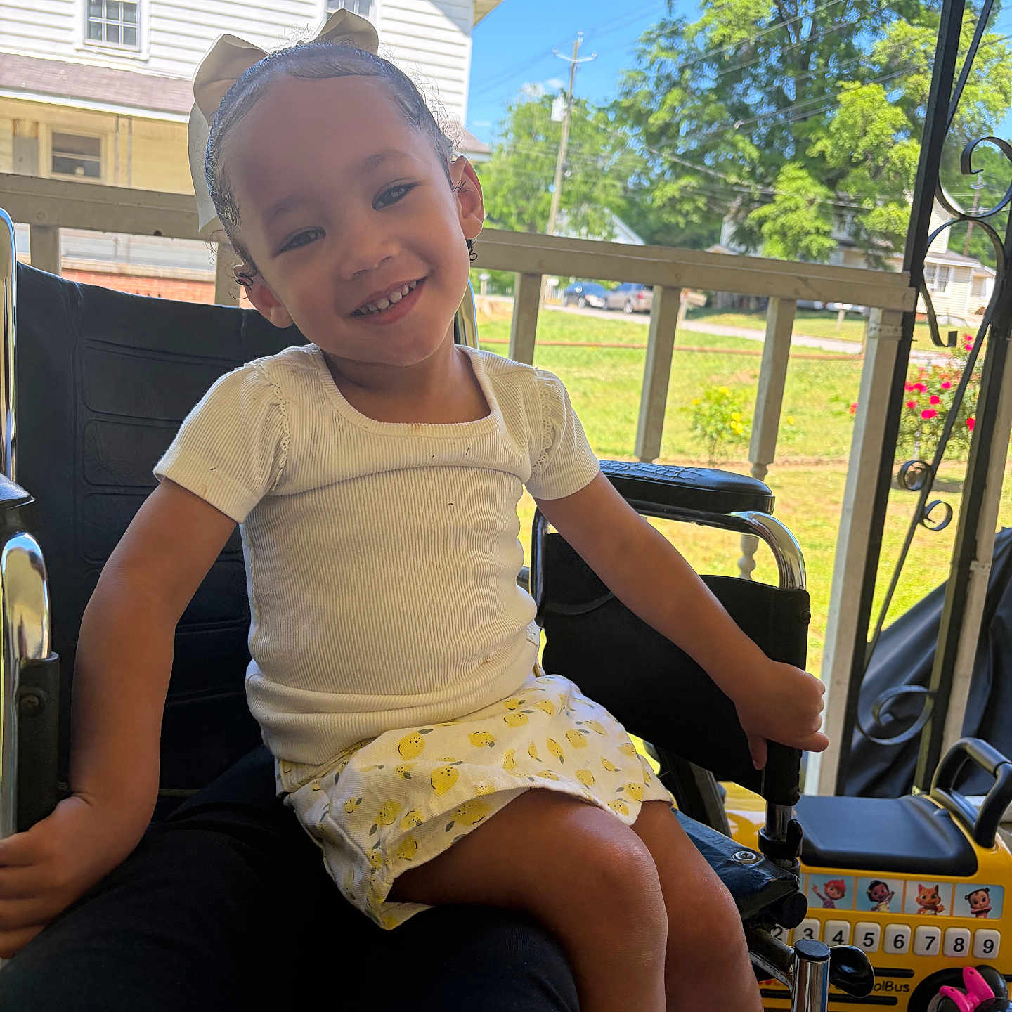 Aneria joined the competition — help win amazing prizes! child, girl, smiling, bow, white_shirt, yellow_skirt, toy, school_bus, porch, chair, outdoor, daylight, greenery, tree, house, fence, happy, person, legs, hand