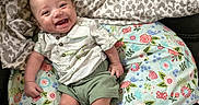 Miles is registered to the contest to win money with this photo: baby, smiling, infant, cushion, blanket, floral_pattern, leopard_print, clothing, green_shorts, short_sleeve_shirt, happy, face, person, indoors, sitting, cute, skin, head, expression, comfort