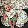 baby, smiling, infant, cushion, blanket, floral_pattern, leopard_print, clothing, green_shorts, short_sleeve_shirt, happy, face, person, indoors, sitting, cute, skin, head, expression, comfort