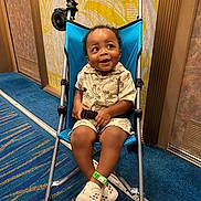 Jaylan is registered to the contest to win money with this photo: toddler, stroller, child, smiling, indoor, blue, clothing, shoes, carpet, patterned_wall, seatbelt, happy, person, footwear, baby, toy_fan, fabric, metal_frame, hand, expression