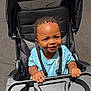 Jaylan is registered to the contest to win money with this photo: toddler, child, stroller, smile, curly_hair, blue_shirt, sunlight, outdoor, tray, hands, seatbelt, pavement, baby, happy, person, infant, cute, young_child, sunny, portrait