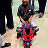 child, toy_motorcycle, indoor, floor, sneakers, jeans, black_shirt, necklace, smile, person, legs, playful, young, casual_clothing, wristband, spiderman_theme, colorful, happy, small_vehicle, people