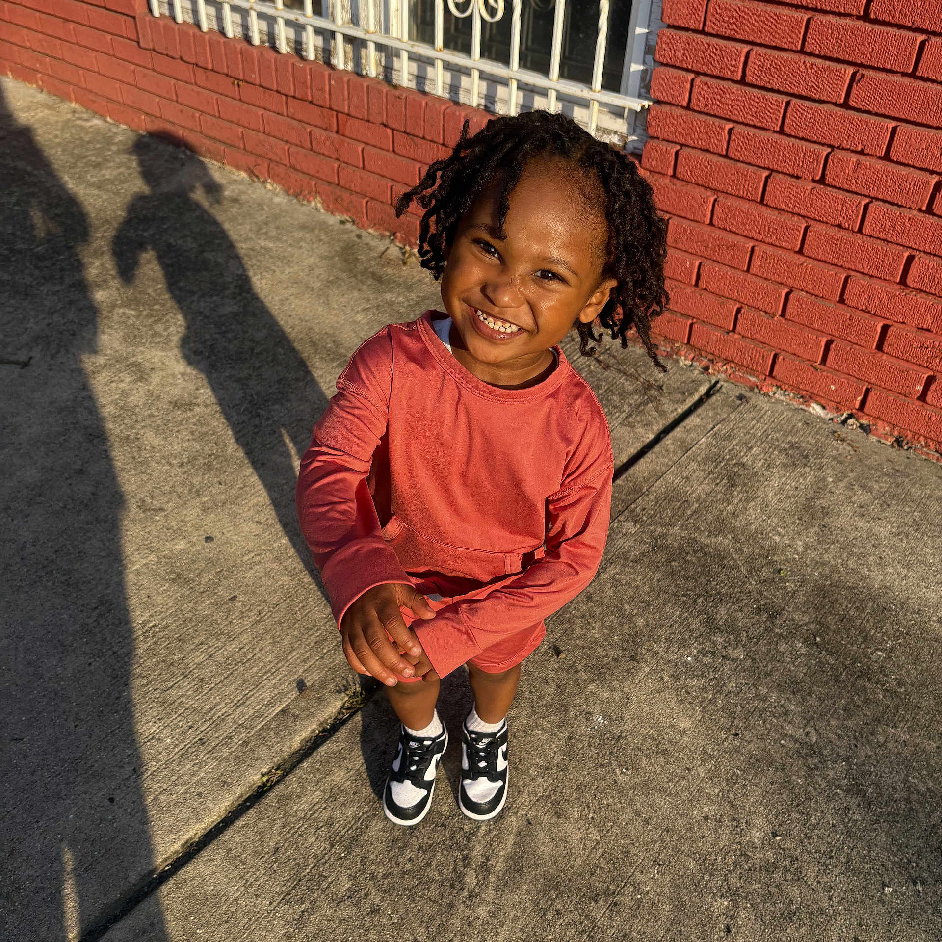 Syeir joined the competition — help win amazing prizes! child, smile, curly_hair, red_brick_wall, sidewalk, shadow, window, sunlight, outdoor, sneakers, happy, person, concrete, casual_clothing, daylight, portrait, standing, urban, iron_bars, playful