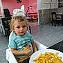toddler, child, high_chair, french_fries, nuggets, plate, food, curly_hair, indoor, table, chair, person, looking, casual_clothing, blue_shirt, yellow_pants, floor, foosball_table, dining_area, modern_interior