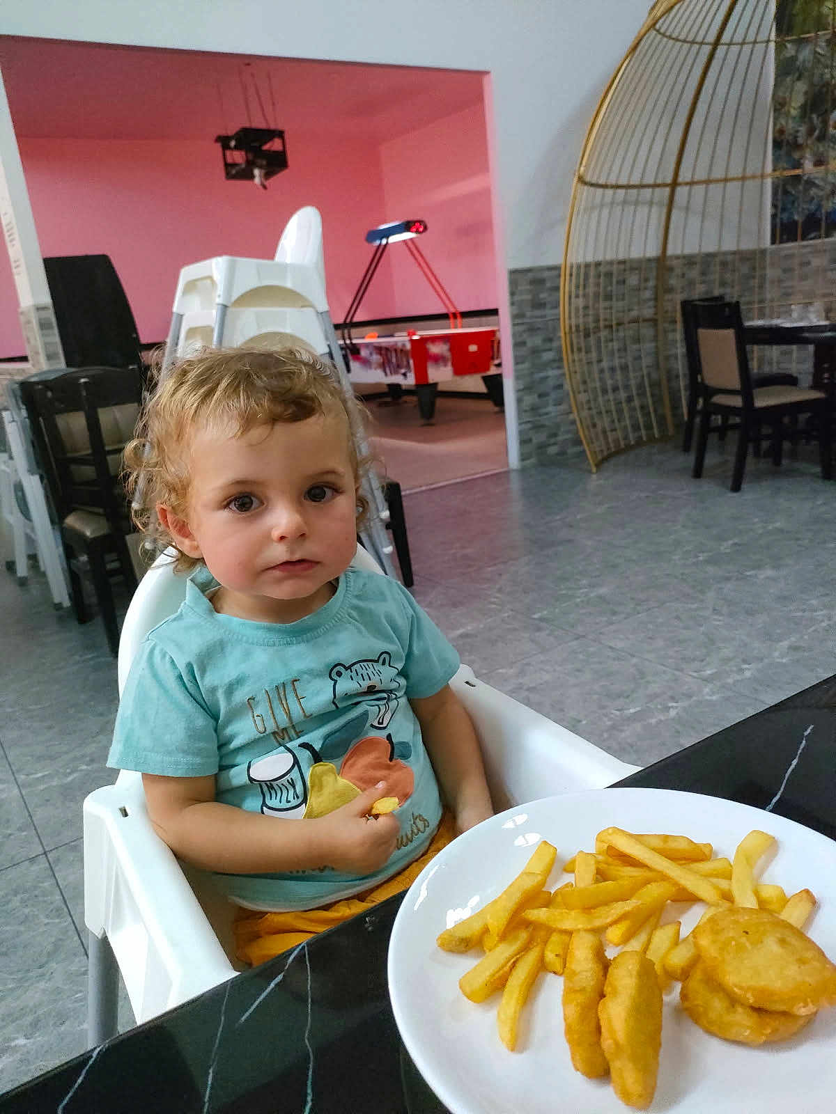 Francis participe au concours pour gagner de l'argent avec cette photo : toddler, child, high_chair, french_fries, nuggets, plate, food, curly_hair, indoor, table, chair, person, looking, casual_clothing, blue_shirt, yellow_pants, floor, foosball_table, dining_area, modern_interior