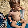 toddler, child, beach, towel, snack, container, barefoot, sand, rocks, bag, shoes, sunlight, outdoor, curly_hair, summer, sitting, casual, playful, daytime, relaxed